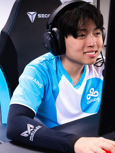 Person wearing a gaming chair with a logo, using headphones and a mouse, in front of a computer screen.